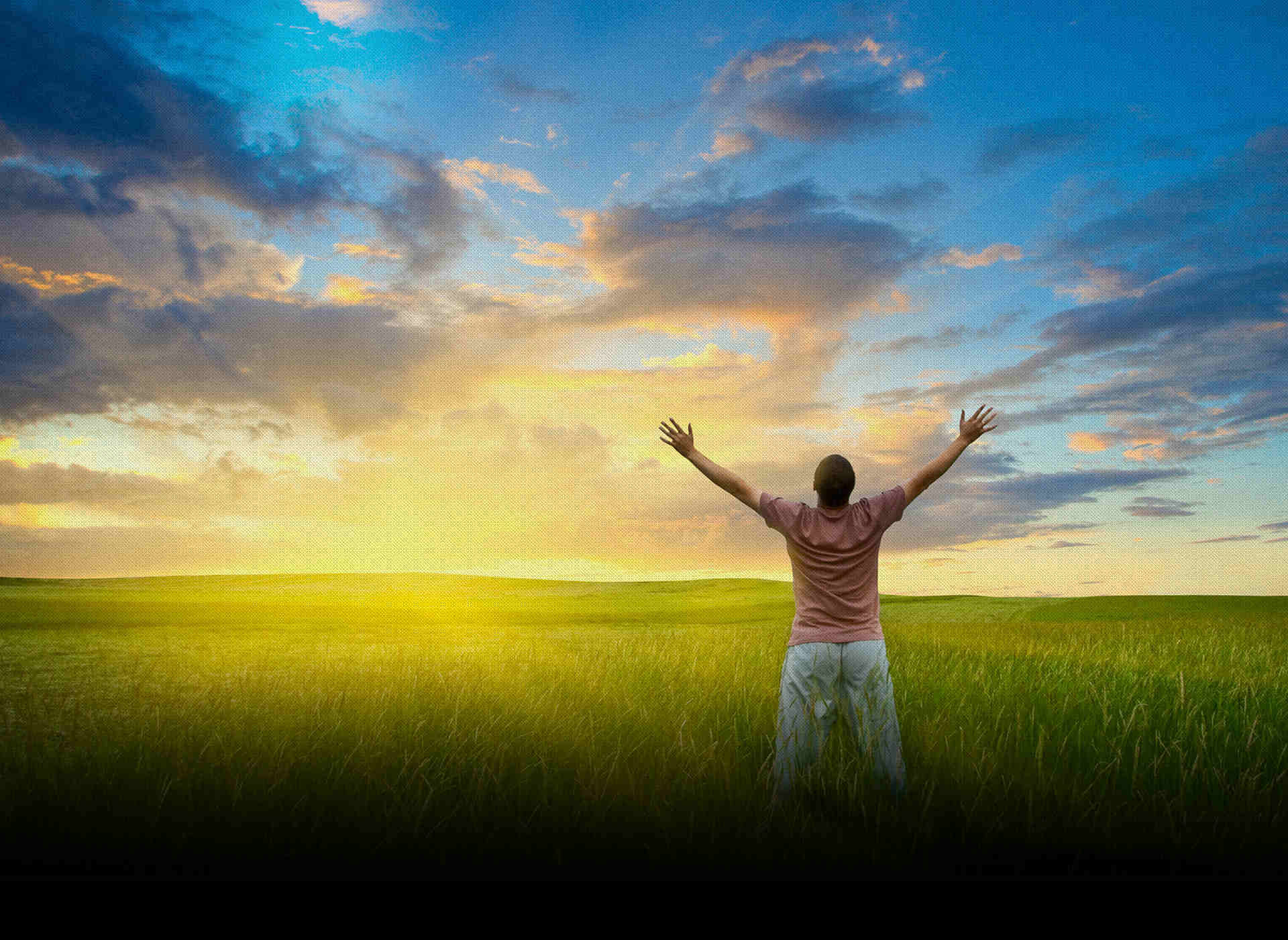 Silhouette of a person with arms raised in a green field under a dramatic sunset sky in blue, yellow and orange tones, adventurous travel mood