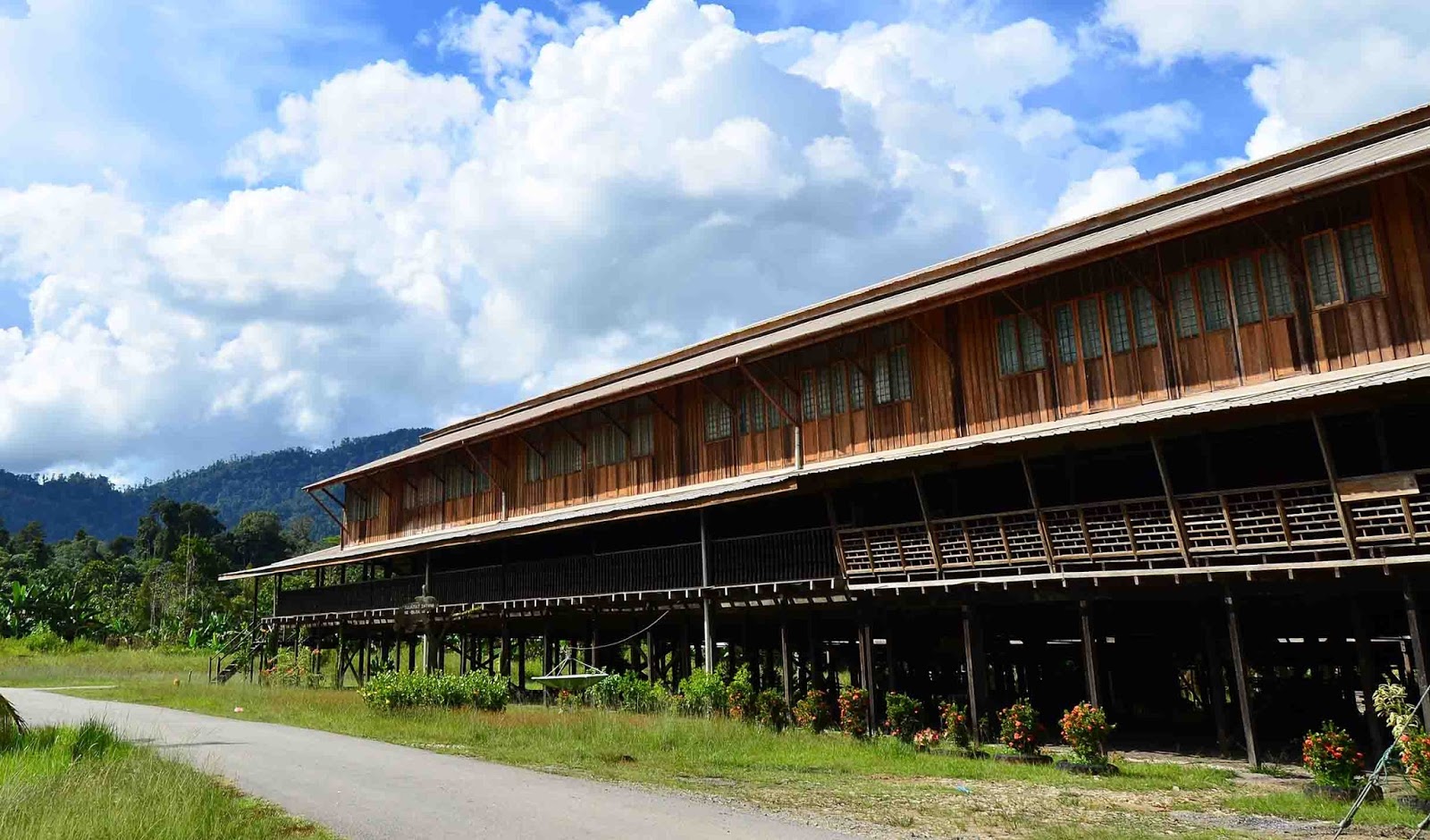Long traditional wooden longhouse on stilts surrounded by greenery and mountains under a blue sky with clouds, calm and earthy travel mood